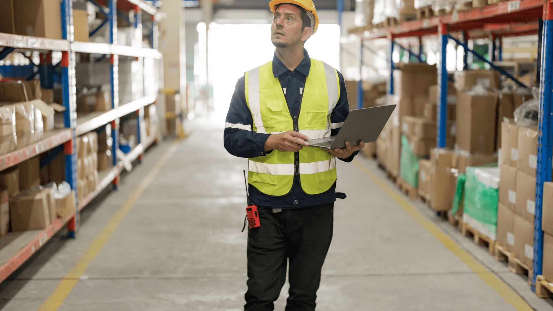 Warehouse worker in a hard hat and safety vest holding a laptop walks between shelves stacked with boxes.