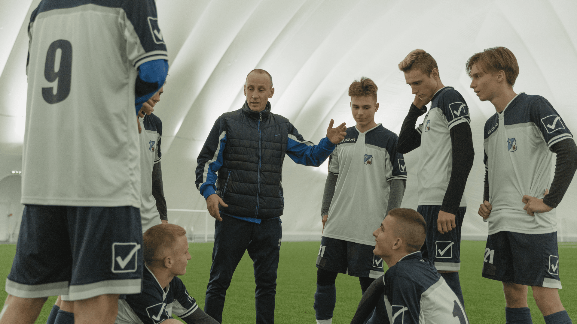 A soccer coach instructs a group of young players in a huddle during an indoor practice.