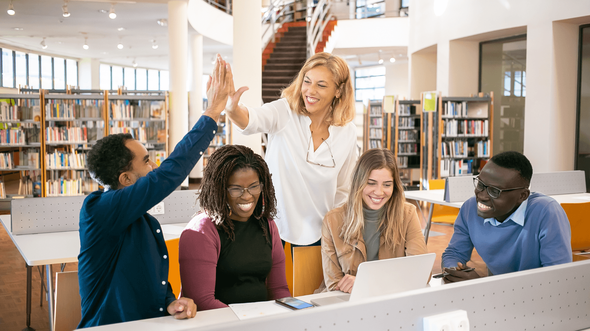 A group of diverse students and a teacher celebrate a successful collaboration in a library setting.