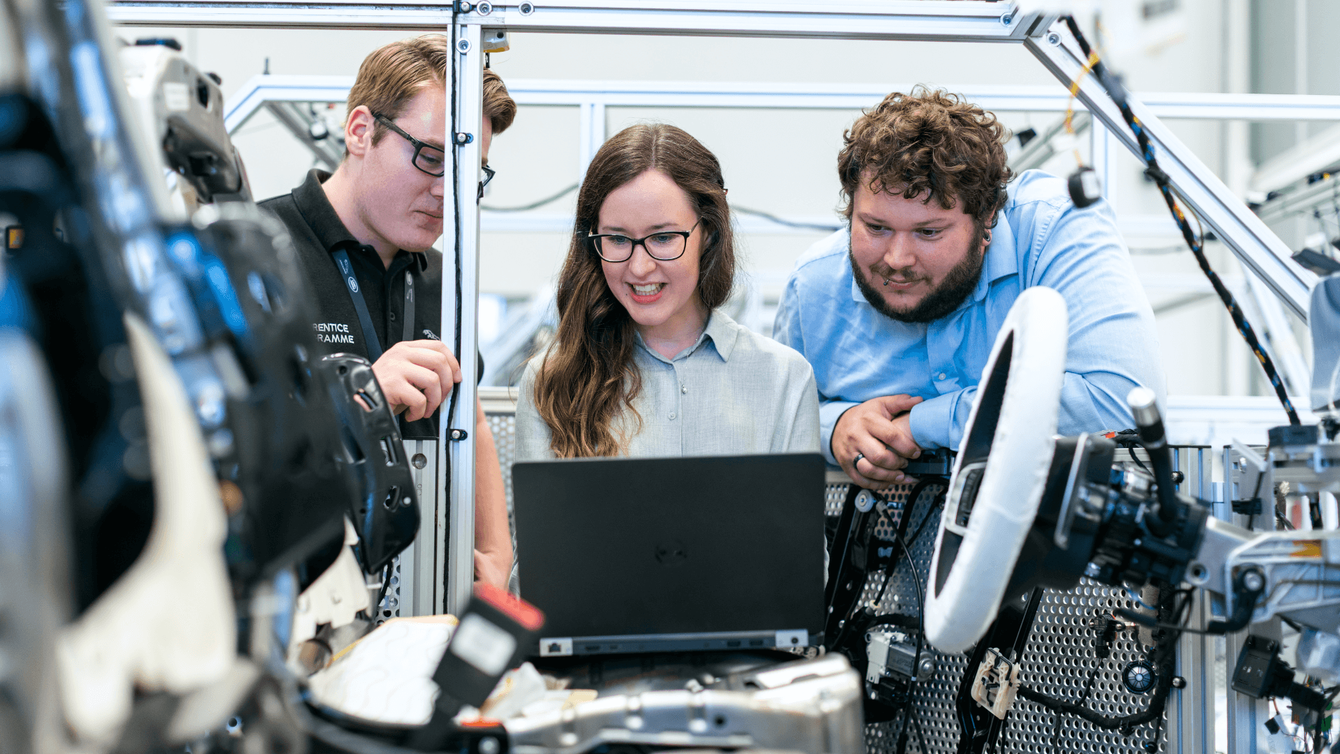Three engineers, two men and one woman, huddle around a laptop in a workshop setting with visible machinery.
