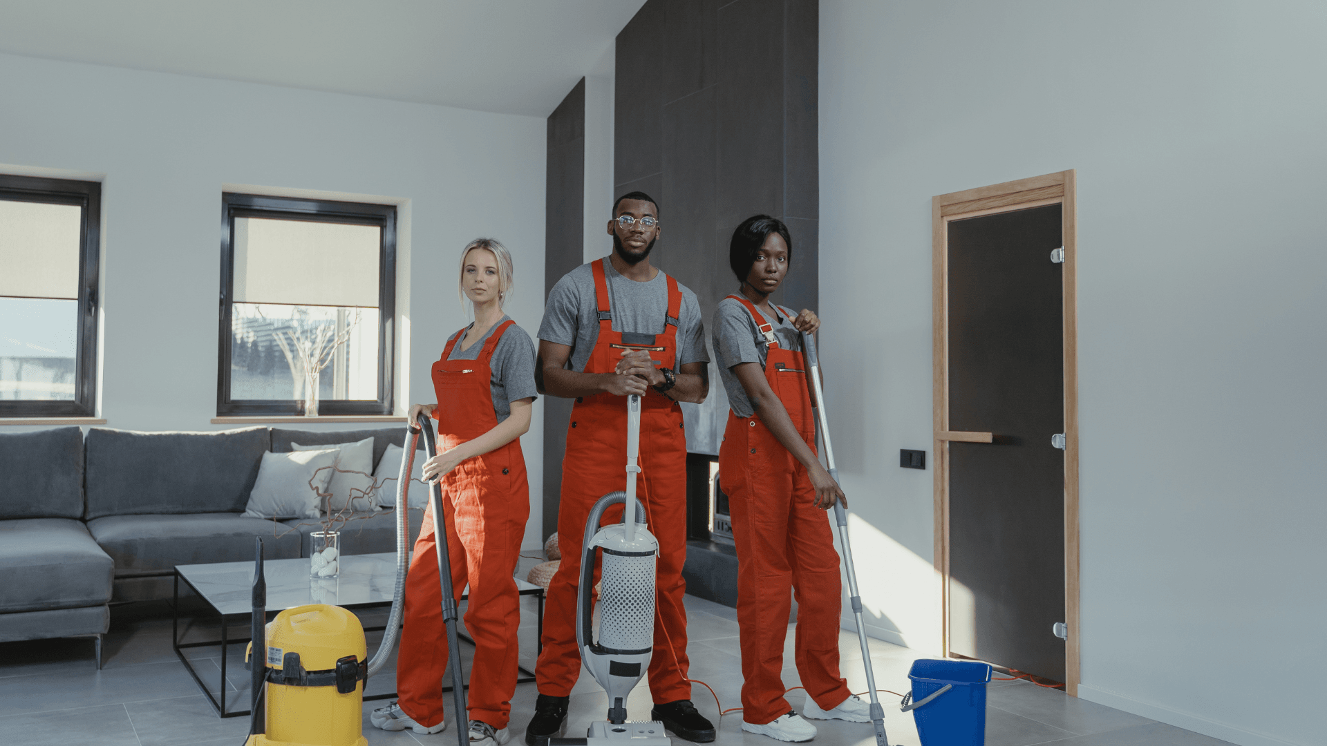 Three cleaning professionals in orange overalls stand with vacuums and mops in a modern living room.
